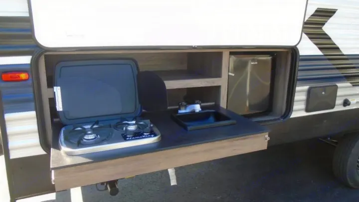 An RV rental's pull-out outdoor kitchen: two-burner stove, sink, mini-fridge, and storage shelves ready for use.. Crossroads RV Zinger 2022