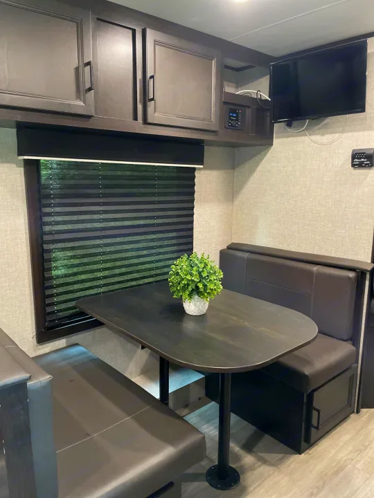 Inside an RV rental: a modern dinette with dark benches, table, window blinds, and overhead storage with TV.. Jayco Jay Flight 2022