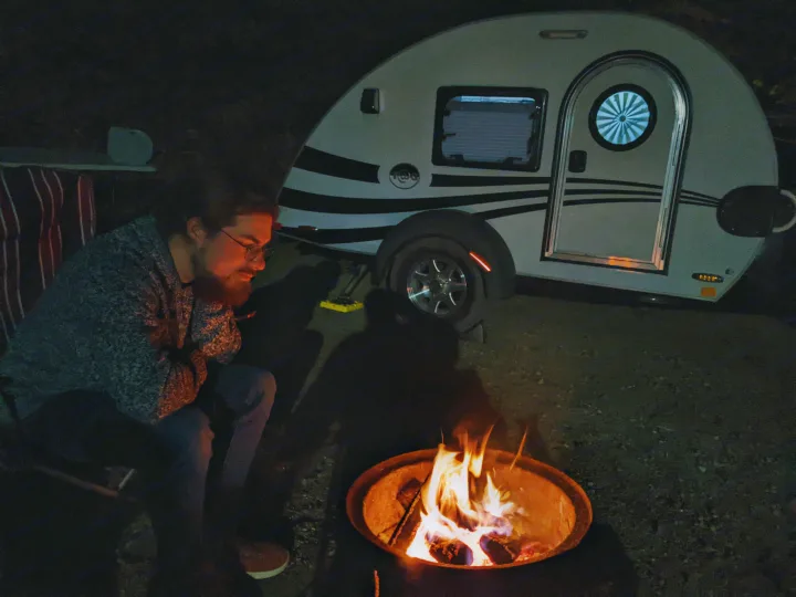 Man by a cozy campfire with a white teardrop RV rental in the dark, enjoying a peaceful camping night.. T@G XL 2016