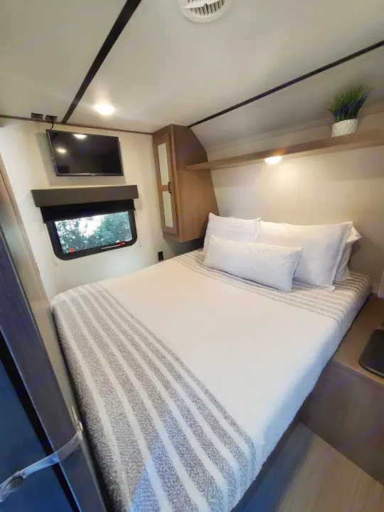 This cozy RV rental bedroom features a comfy bed, wall-mounted TV, window, and overhead storage.. Keystone RV Hideout Luxury 2021