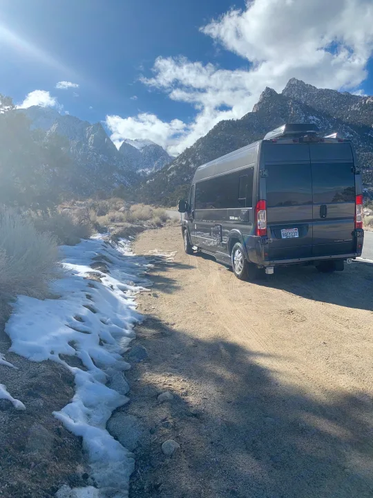 Scenic RV rental journey: Gray van on a dirt road with snow-dusted mountains & blue sky. Bright sun, perfect for adventure.. Roadtrek Roadtrek Class B 2018