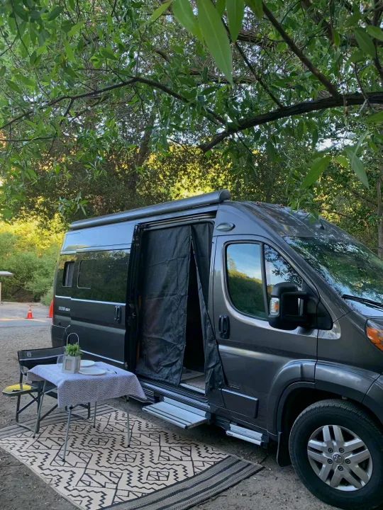 Cozy RV rental setup with a gray camper van, outdoor rug, and dining table, nestled in a serene natural setting.. Roadtrek Roadtrek Class B 2018