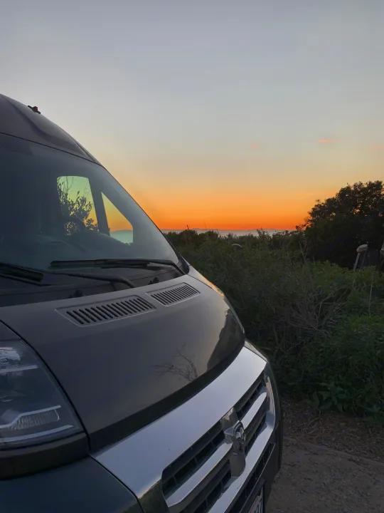 RV rental van parked, showcasing a vibrant orange and blue sunset over the ocean and coastal bushes.. Roadtrek Roadtrek Class B 2018