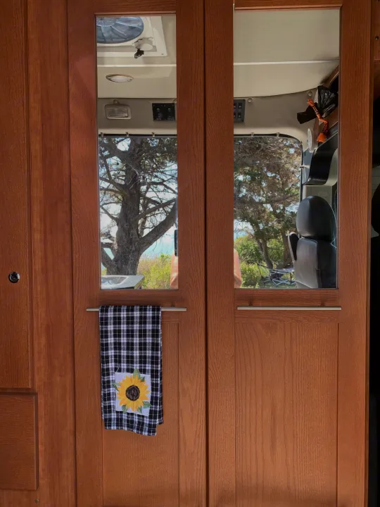 Inside an RV rental, showing warm wood doors with glass panels revealing trees and distant water outside. A sunflower towel hangs.. Roadtrek Roadtrek Class B 2018