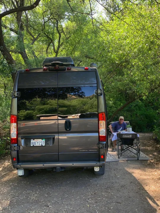 Outdoor RV rental setup: a man relaxes at a table beside his gray camper van surrounded by green trees.. Roadtrek Roadtrek Class B 2018