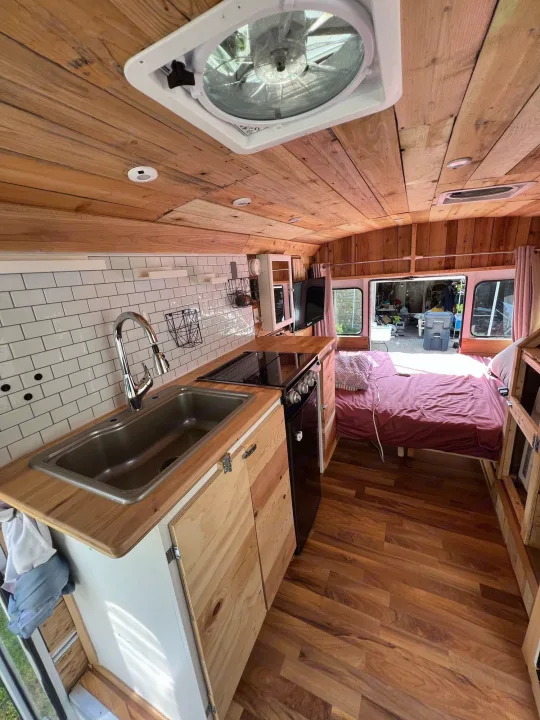 Custom RV rental interior featuring a wood-paneled ceiling & walls, full kitchen with sink & stove, and a cozy bed area.. Chevy Express 3500 Mini Bus 2002