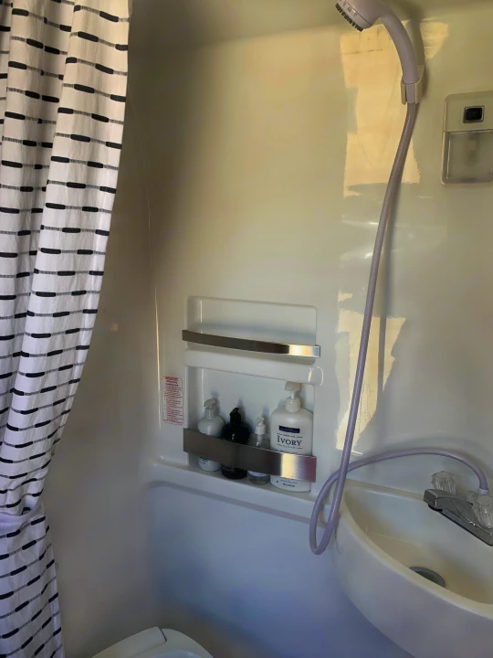 Inside a bright white RV rental bathroom, featuring a shower head, sink, and striped shower curtain. Toiletries are on a built-in shelf.. Roadtrek Roadtrek Class B 2018