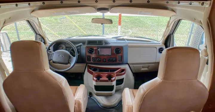 Inside an RV rental: a view of the front cockpit from behind the seats, showing the Ford dashboard, steering wheel, and console.. Coachman Leprechaun 260DS 2017