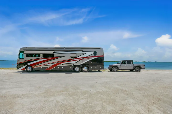 RV rental adventure: A large motorhome towing a Jeep Gladiator on a sandy beach by clear blue water.. Ventana Ventana Motorhome 2022