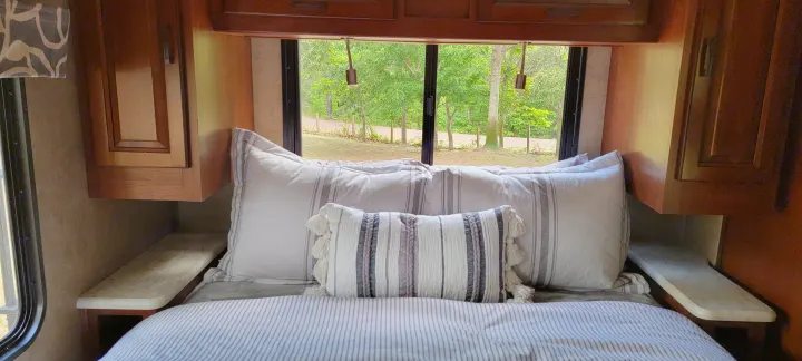 Cozy RV rental bedroom featuring a comfortable bed with striped linens and a large window overlooking a lush, wooded path.. Forest River 3171DS 2016