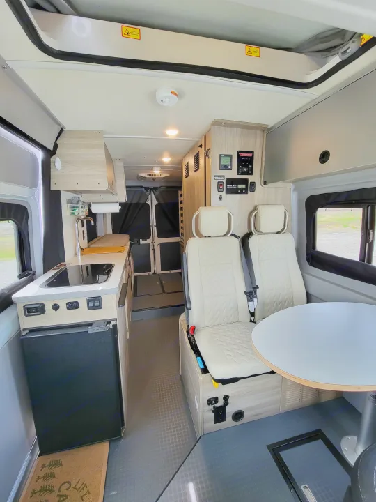Inside a bright rv rental, showing front-facing passenger seats with a table, control panel, and integrated kitchenette at the rear.. Winnebago Solis 2021