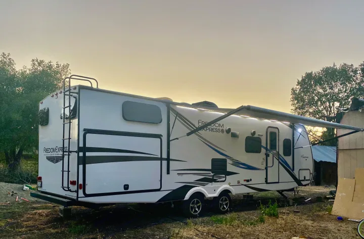 A white Freedom Express travel trailer with its lighted awning extended. Perfect for an RV rental and outdoor adventures.. Coachmen Freedom 2021