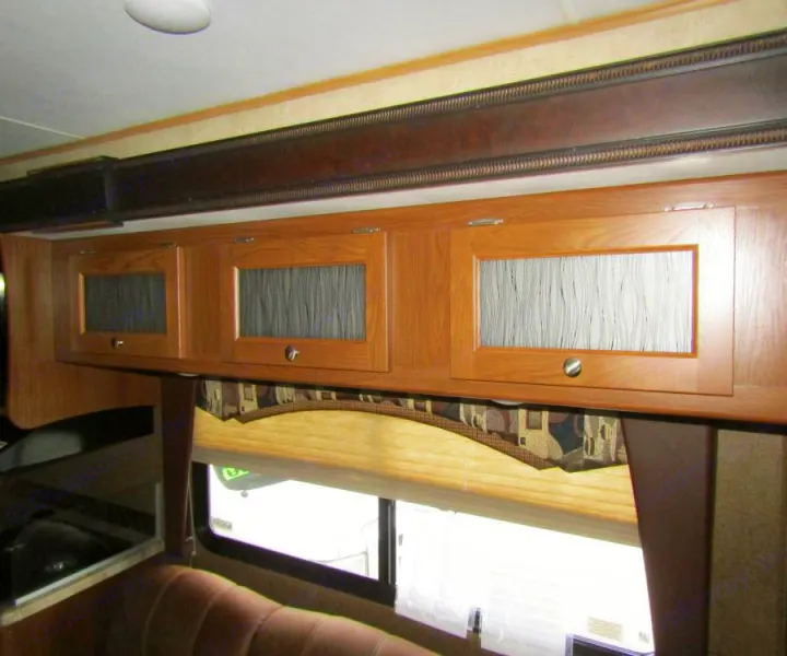 Inside an RV rental: wood overhead storage cabinets with glass inserts, window with shade, & seating.. Coachmen Leprechaun 2017