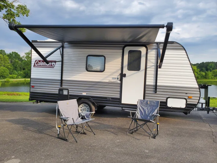 An RV rental travel trailer with an extended awning and camping chairs by a scenic pond.. Coleman LANTERN LT 17B 2022
