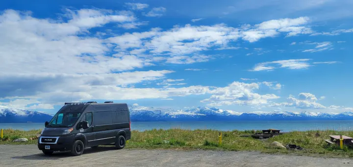 Dark grey RV rental van at a scenic waterfront campsite, with snow-capped mountains, a distant ship, and bright blue sky.. Ram Promaster 2020