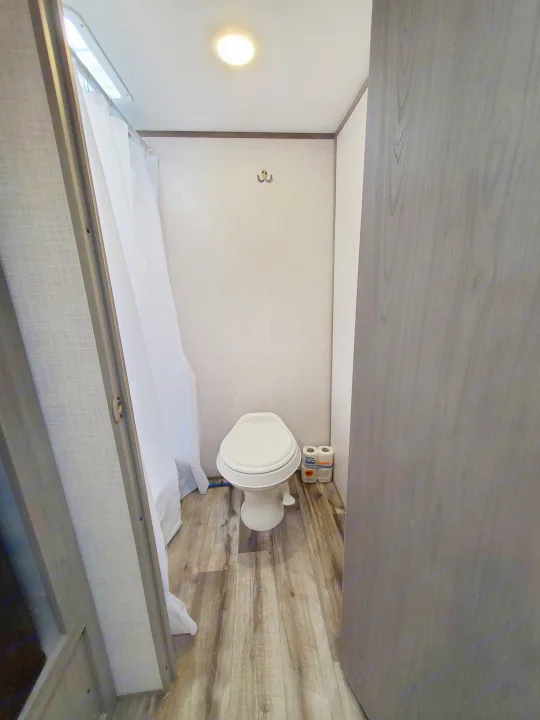 Compact RV rental bathroom: features a toilet, shower curtain on left, wood-look floor, ceiling light, and wall hook.. Coleman LANTERN LT 17B 2022