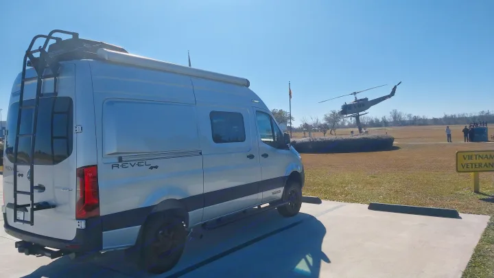 A Mercedes-Benz Sprinter RV rental van (Winnebago Revel 4x4) parked facing a Vietnam veteran helicopter memorial under a clear sky.. Winnebago Revel 2021