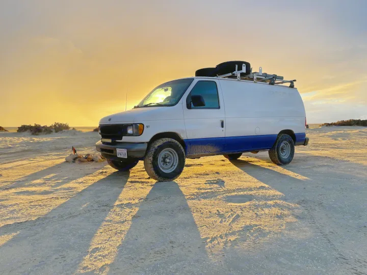 Customized white and blue adventure van (potential RV rental) with off-road tires, parked in sandy terrain at sunset, casting long shadows.. Ford Econolline 250s 2001