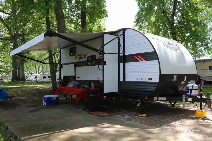 This white and grey Forest River Wildwood FSX travel trailer RV rental is set up in a wooded campground, ready for outdoor living.. Forest River Wildwood FSX 2021