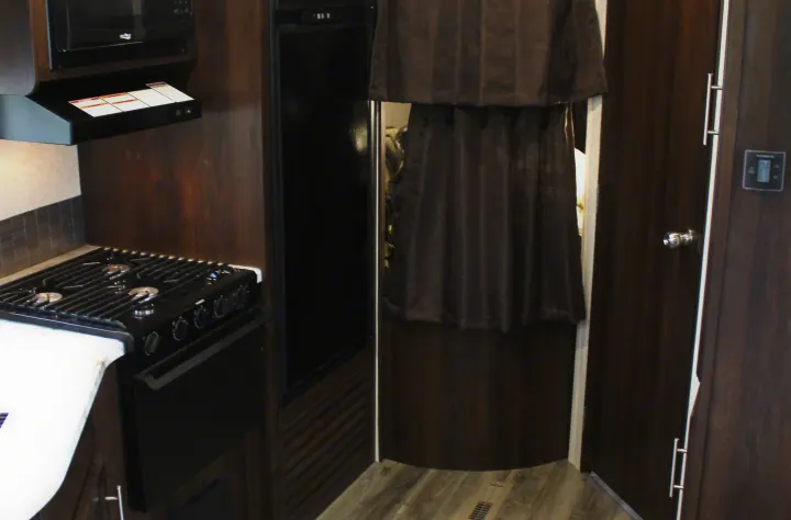 RV rental interior: compact kitchen with stove, microwave, and refrigerator next to a dark curtained doorway.. Jayco Jay Flight SLX Rocky Mtn Edition 2019