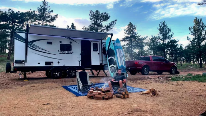 Relaxing campsite scene: RV rental set up with extended awning, truck, man by campfire, paddleboards, and a dog.. Heartland RVs North Trail 2021