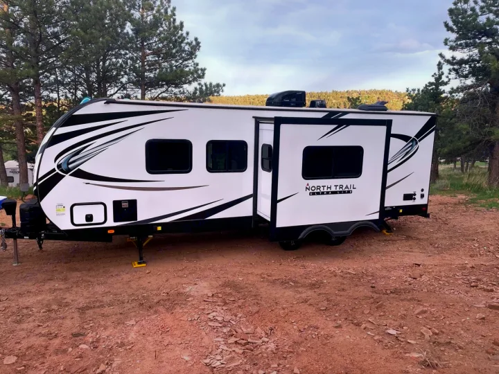 White North Trail travel trailer RV rental, with extended slide, nestled among pine trees on red dirt.. Heartland RVs North Trail 2021