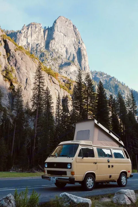 A tan VW Westfalia camper van, ideal for an RV rental, parked on a scenic road with majestic granite mountains and tall pine forests.. Volkswagen Westfalia 1982