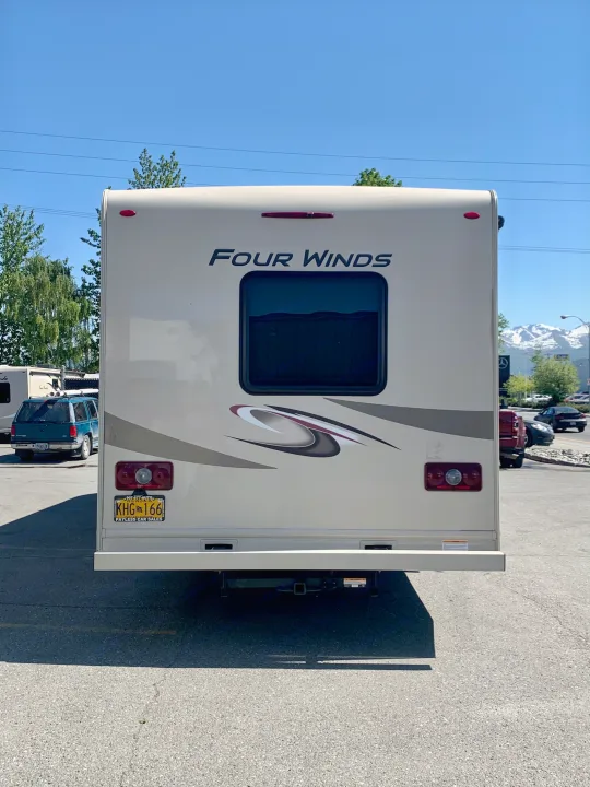 A beige Four Winds RV rental, seen from the rear, with Alaska plate KHG 166, showing its tow hitch and distant snowy mountains.. Thor Motor Coach Four Winds 2019