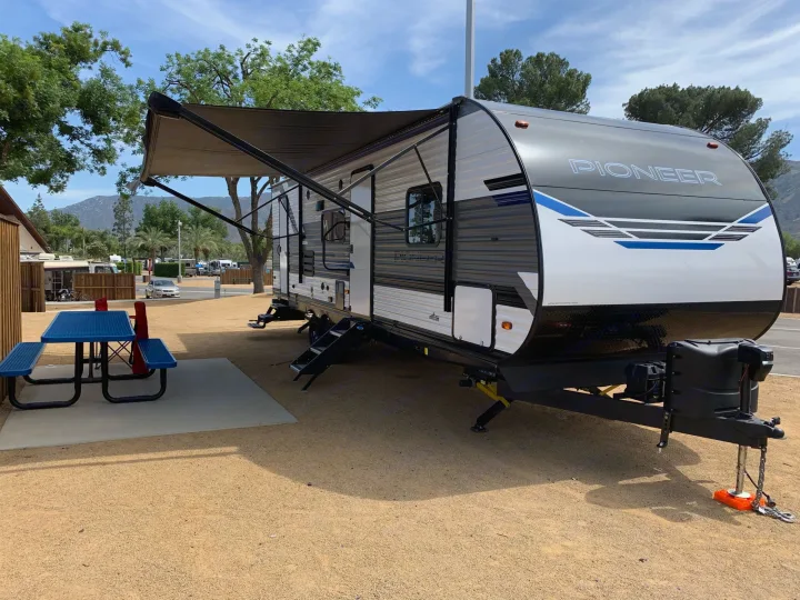 A modern Pioneer travel trailer RV rental with an extended awning and a blue picnic table at a sunny, scenic campsite.. Heartland Pioneer 2022