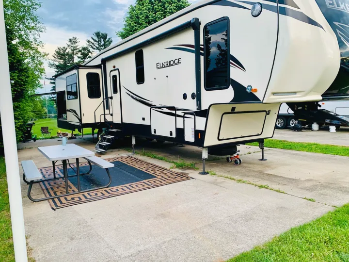 A spacious ELKRIDGE fifth-wheel RV rental is set up at a concrete campsite, complete with an awning, picnic table, and outdoor rug.. Heartland Elkridge 2019