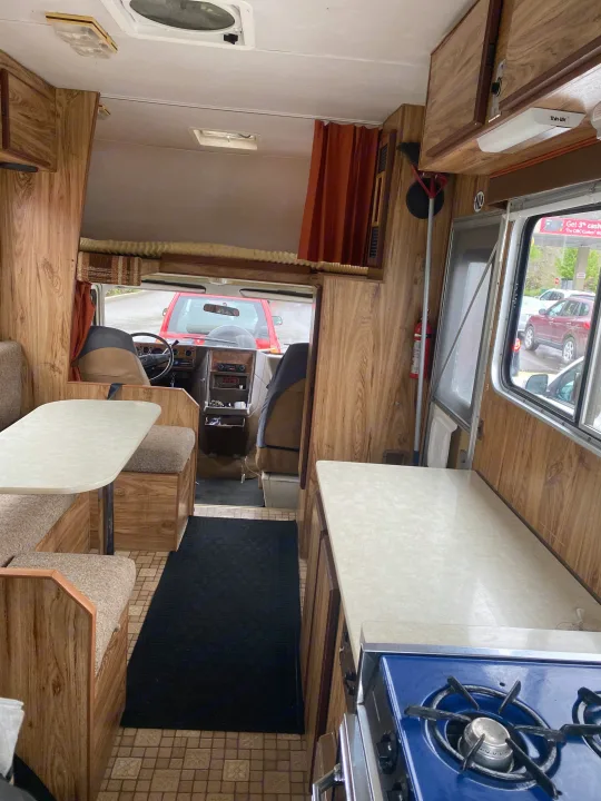Experience a retro RV rental interior! Features include a dinette, blue stove in the kitchen, and warm wood paneling throughout.. Vanguard Industries MV 1800 1981