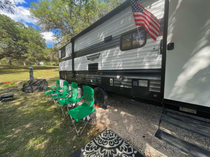 Relaxing RV rental campsite. Features travel trailer with US flag, green folding chairs, and a Blackstone grill.. Forest River Wildwood X-Lite 2021