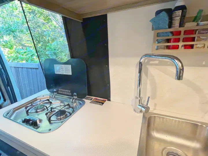 Compact RV rental kitchen features a two-burner stove, sink, and ample counter space, with a leafy outdoor view.. Winnebago Winnebago Solis 59P 2022