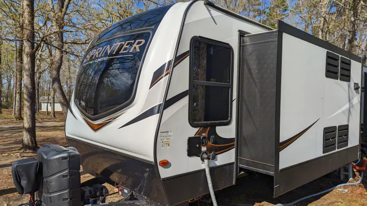 A white Sprinter travel trailer, perfect for rv rental, parked in a sunny, wooded campsite. Features black/gold accents.. Keystone RV Sprinter Limited 2021