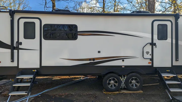 Full side view of a white travel trailer, an RV rental, showing doors with steps, wheels, and an awning. Parked outdoors.. Keystone RV Sprinter Limited 2021