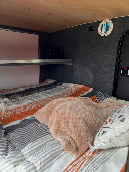 An inviting rv rental sleeping area, featuring a striped bedspread, soft blanket, wooden ceiling, and a practical shelf unit.. Sherpa trailers Big foot 2022