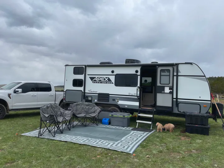RV rental setup: White truck & Coachmen Apex travel trailer on grass. Outdoor mat, chairs, & two fluffy dogs complete the camp.. Coachmen Apex 2022