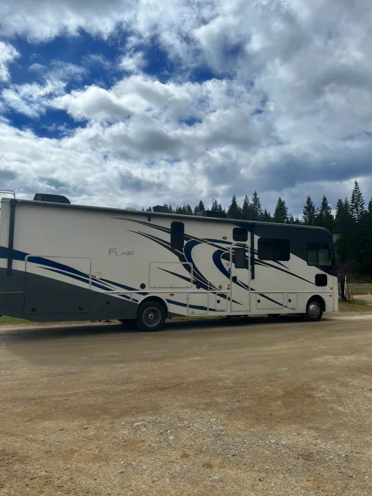 White Flair RV rental on a gravel road, with pine trees and cloudy sky. Perfect for outdoor adventures.. Flair-Fleetwood Flair-Fleetwood Motorhome 2020