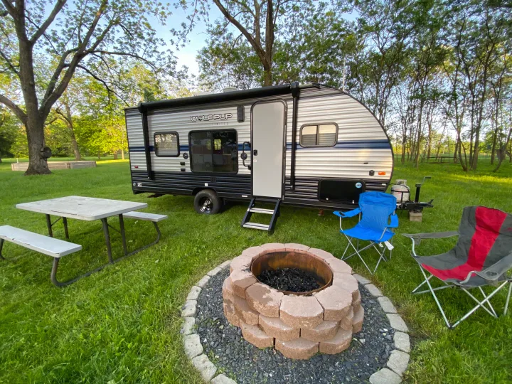 A 'Wolf Pup' RV rental is parked on green grass next to a picnic table, fire pit, and two chairs under trees.. Forest River Cherokee Wolf Pup 2019