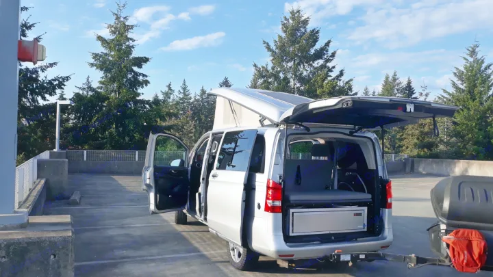 Silver pop-top RV rental camper van with open doors and a cargo box. Parked in a concrete lot amidst green trees.. Mercedes Metris 2016