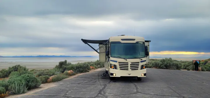 An RV rental (Class A motorhome) by a vast lake & mountains under a cloudy sky. Awning extended; person by sign.. FR3 FR3 Motorhome 2020