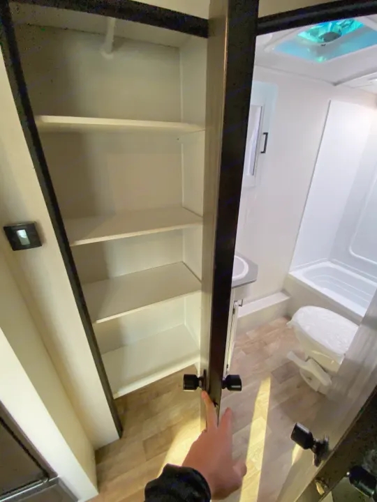 Inside an rv rental, a hand opens a tall, shelved cabinet in a compact bathroom with a toilet, sink, and shower.. Keystone RV Hideout 2022