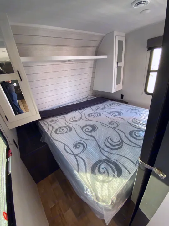 Compact RV rental bedroom with new, plastic-covered mattress, overhead cabinet, and wall shelving.. Keystone RV Hideout 2022