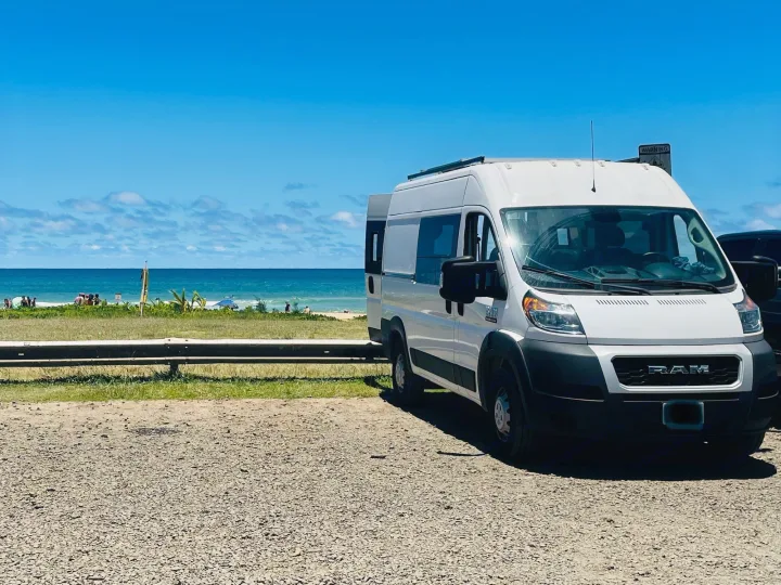 White Ram ProMaster RV rental van on gravel, facing a beautiful blue beach with ocean under a clear sky.. Dodge Ram Promaster 1500 2021