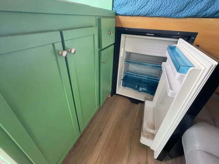 Compact RV rental kitchen featuring green cabinets, wood floors, and an open built-in refrigerator with blue shelves.. Dodge Ram Promaster 1500 2021