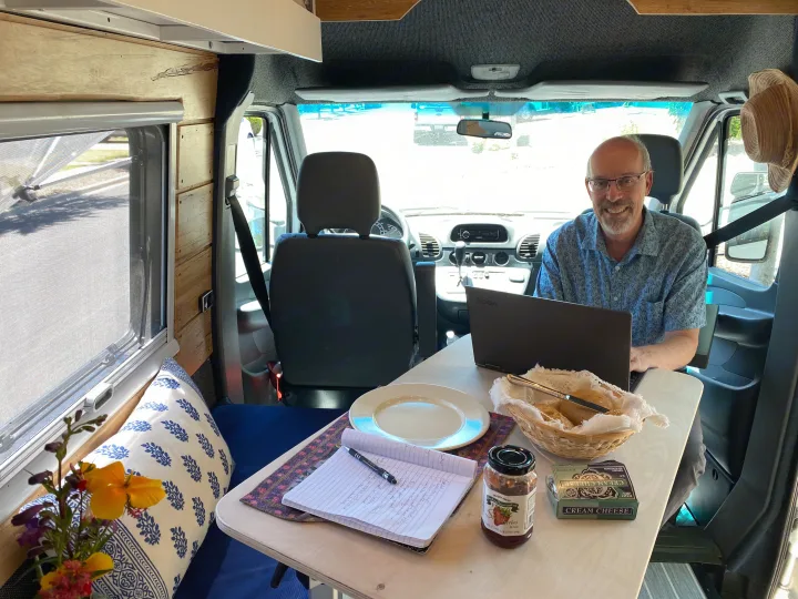 A man works on his laptop inside a cozy RV rental, with snacks and a meal ready at his mobile workstation.. Mercedes Sprinter 2005