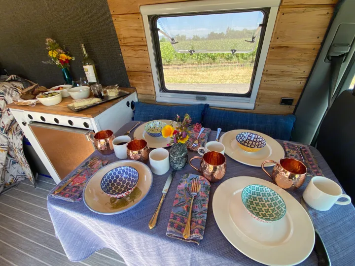 A cozy RV rental interior with a table set for a meal, featuring fruit, cheese, wine, and a scenic vineyard view outside.. Mercedes Sprinter 2005