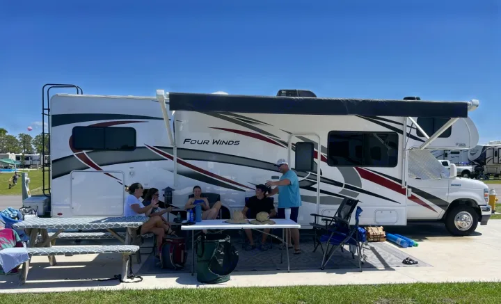 A group of people enjoys a sunny day at their RV rental campsite, gathered under the awning with chairs and a table.. Thor Four Winds 2022
