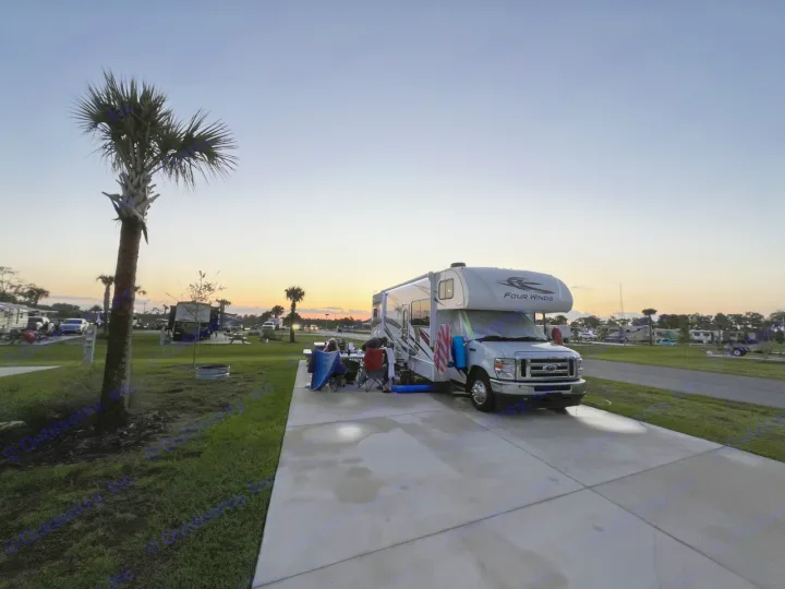 "Four Winds" RV rental at a palm-lined campground at sunset. People relax outside.. Thor Four Winds 2022