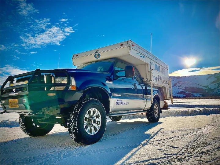 Blue Ford truck with Arctic Overland camper on snow. Heart-shaped sun. Perfect RV rental for Alaska winter trips with your dog.. Ford F250 2005
